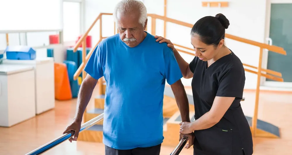 Physiotherapist supporting an older man as he walks between parallel bars